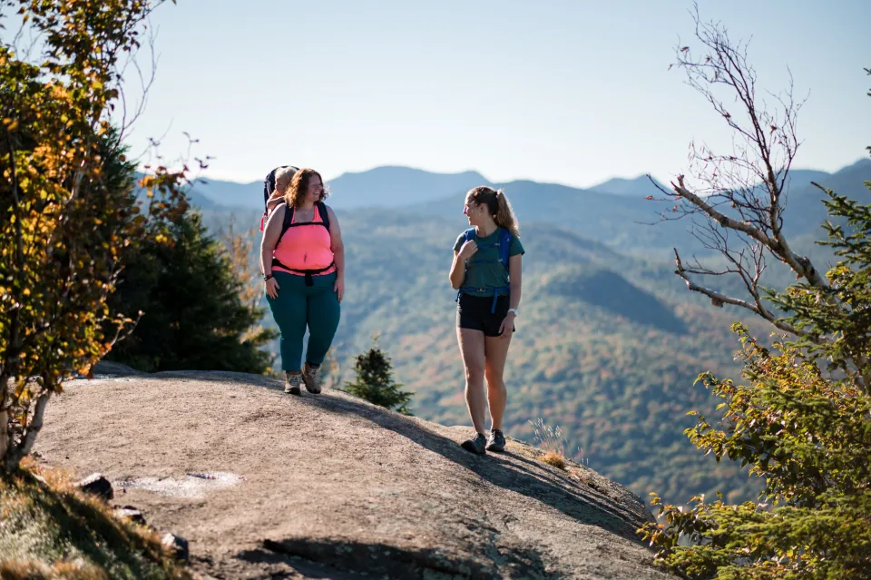 Two women, one with a baby carrier, stand atop a mountain summit